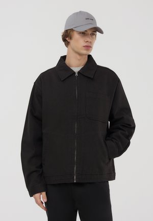Giacca in denim nera con zip frontale, colletto appuntito e una tasca sul petto. Il modello indossa un cappellino da baseball grigio chiaro, posizionato su uno sfondo bianco.