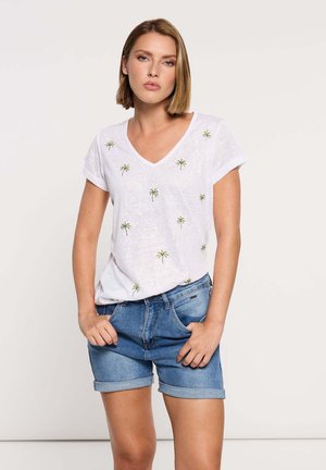 Mujer con cabello rubio corto, vestida con una camiseta blanca de cuello en V con pequeños estampados de palmeras y shorts de mezclilla remangados, con las manos en los bolsillos.