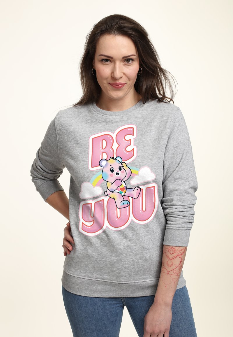Sudadera gris con un gráfico colorido de un oso de dibujos animados, con el texto "SÉ TÚ MISMO" en rosa y amarillo, y acentos de arcoíris y nubes.