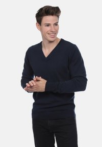 Jeune homme souriant, portant un pull en V bleu marine et un pantalon noir, les mains croisées et regardant à sa gauche.