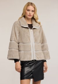 Blondýnka v béžovém umělém kožichu s širokými rukávy a asymetrické černé kožené sukni, stojí na jednoduchém pozadí.