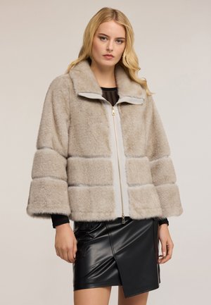 Blonde Frau, die eine beige Kunstfelljacke mit breiten Ärmeln und einen asymmetrischen schwarzen Lederrock trägt, steht vor einem einfarbigen Hintergrund.