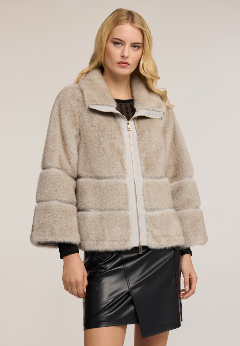 Blondýnka v béžovém umělém kožichu s širokými rukávy a asymetrické černé kožené sukni, stojí na jednoduchém pozadí.