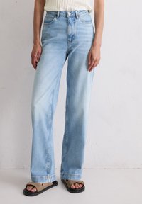 Jasnoniebieskie dżinsy z wysokim stanem o luźnym kroju, z pięcioma kieszeniami i delikatnym wyblaknięciem. Połączone z czarnymi i beżowymi sandałami.