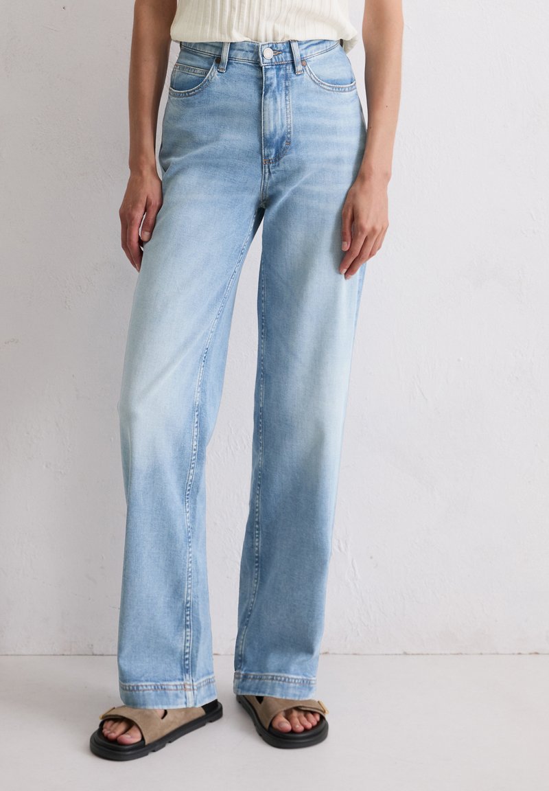 Jasnoniebieskie dżinsy z wysokim stanem o luźnym kroju, z pięcioma kieszeniami i delikatnym wyblaknięciem. Połączone z czarnymi i beżowymi sandałami.