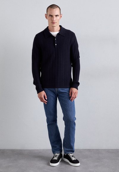 Polo din tricot texturat, de culoare bleumarin, cu guler deschis, asortat cu blugi drepti de un albastru deschis și adidași negri și albi.