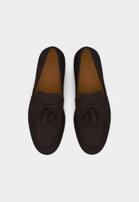 Paire de mocassins en daim brun foncé avec pompons, couture style mocassin et semelles intérieures en cuir beige, vue de dessus sur fond blanc.