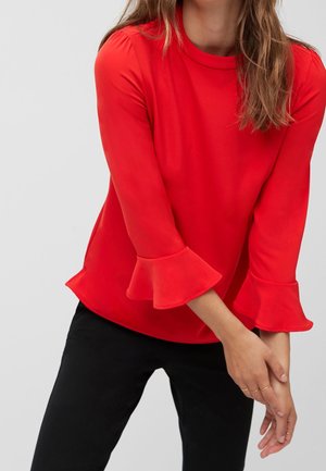 Femme portant un chemisier rouge vif à manches longues avec des poignets volantés et un pantalon noir, debout devant un fond uni gris clair.