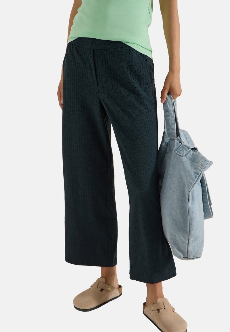 Personne portant un haut vert clair, un pantalon noir ample, des chaussures beiges à enfiler, tenant un sac en jean bleu clair, montrée des épaules vers le bas.
