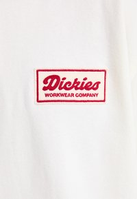 Hvit bomullsstoff har et rødt brodert merke med "Dickies" og "WORKWEAR COMPANY" i fet skrift på en kremfarget bakgrunn.