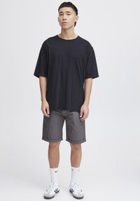 Schwarzes, locker sitzendes T-Shirt, graue Jeansshorts mit umgeschlagenen Säumen, weiße Socken und schwarz-weiße Turnschuhe. Schlichte, legere Kleidung.