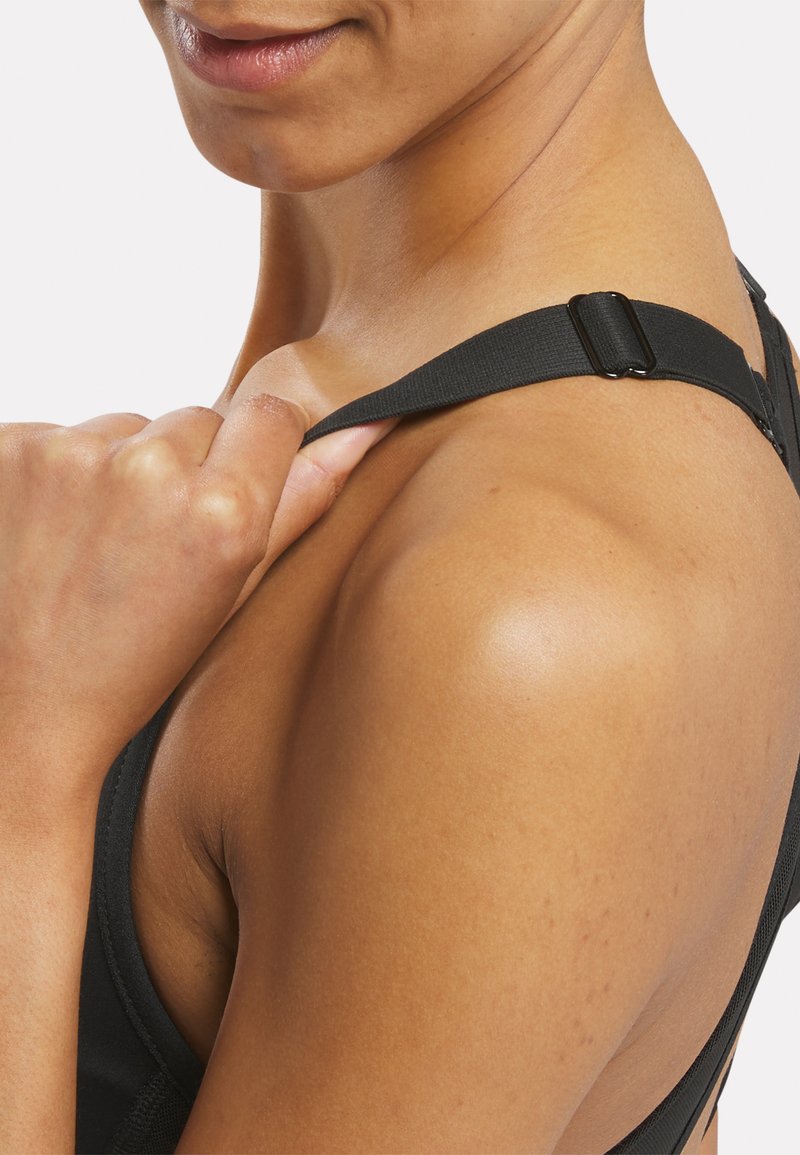Tirante de sujetador deportivo negro con herrajes ajustables; textura suave, banda ancha y un primer plano de la piel que revela un tono uniforme y ligeras pecas.