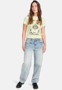 Light yellow graphic t-shirt with an eye design, paired with faded blue loose-fitting denim jeans and black shoes. Casual outfit.