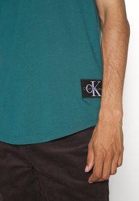 Türkisfarbenes Baumwoll-T-Shirt mit abgerundetem Saum. Mit einem schwarzen Stoffetikett mit weißem "CK"-Logo. Darunter werden dunkelbraune Cordhosen getragen.