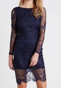 Marinblå spetsklänning med långa genomskinliga ärmar, blommönster och foder. Nederkanten har spetsdekoration och klänningen når till mitten av låren.