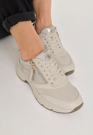Zapatillas de deporte color beige claro con materiales de ante y malla, que presentan un diseño con cordones, acentos plateados y un detalle de cremallera lateral.
