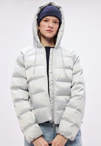 Svijetlosiva jakna s punjenjem, kapuljačom, visokim ovratnikom i elastičnim manžetama; sadrži prednji zip i tuftanu teksturu za izolaciju.