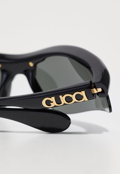 Juodos akiniai nuo saulės su blizgiu paviršiumi, su auksiniu "GUCCI" logotipu ant kojelės ir tamsiai pilkais lęšiais. Suapvalinta, modernaus dizaino.