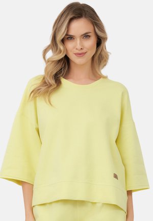 Kobieta z falującymi blond włosami ubrana w luźną, pastelowo żółtą bluzkę z rękawem za łokieć oraz dopasowane spodnie na białym tle.