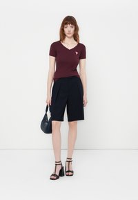 Donna in piedi contro un muro bianco che indossa una maglietta bordeaux a scollo a V, pantaloni corti neri fino al ginocchio, sandali con tacco neri e tiene in mano una borsa nera.