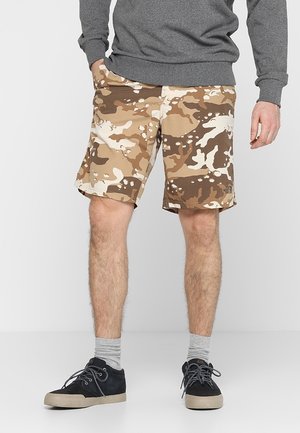Mann trägt beige-braune Tarnshorts, grauen Pullover, graue Socken und schwarze Turnschuhe, steht mit entspannten Händen an den Seiten.