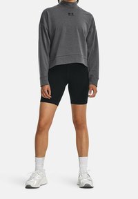 Persoon in een grijze lange mouw cropped sweatshirt, zwarte biker shorts, witte sokken en witte sportschoenen, staand tegen een effen achtergrond.