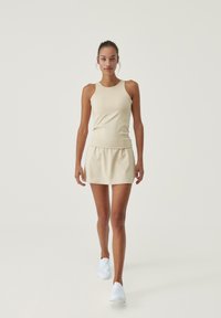 Beige athletic tank top paired with a matching flared skirt, both made of smooth, stretchy fabric; model wears white slip-on shoes.