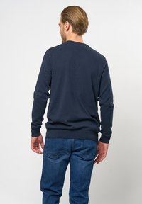Sötétkék pulóver bordázott mandzsettával és hem, kerek nyakkivágással, sima textúrával. Kék farmernadrággal viselve, hátulról nézve.