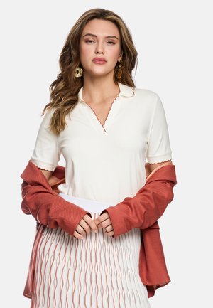 Frau mit welligem Haar trägt goldene Ohrringe, weißes Kurzarmbluse mit Wellensaum, rostfarbenen Jacke, die über den Armen hängt, und gemusterten Rock.