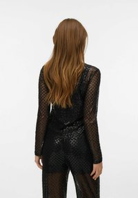 Zwarte jumpsuit met lange mouwen, voorzien van een geometrisch mesh-patroon, semi-transparante textuur en een getailleerd silhouet, gezien vanaf de achterkant.