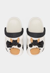 Λευκά παιδικά Crocs με μαύρα ρυθμιζόμενα λουράκια και πρόσωπα αγελάδας που διαθέτουν αυτιά, μάτια και καφέ μουσούδες στην περιοχή των δακτύλων.