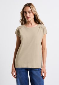 Beige tričko krátký rukáv s kulatým výstřihem, jemná tkanina a decentní nařasení na ramenních švech, v kombinaci s modrými džíny.