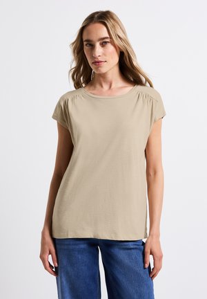 T-shirt beige a maniche corte con scollo rotondo, tessuto morbido e delicate arricciature alle cuciture delle spalle, abbinato a jeans blu.