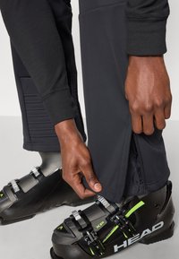 Μαύρο, εφαρμοστό παντελόνι για σκι με φερμουάρ στο κάτω μέρος. Υφασμάτινη επιφάνεια με υφή και ριγωτές μανσέτες. Φορεμένο με μαύρες μπότες σκι που διαθέτουν ρυθμιζόμενα κλιπς.