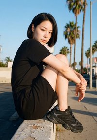 Ung kvinde i sort kjole og sneakers sidder på en betonkant med palmer og klar himmel i en urban setting ved solnedgang.