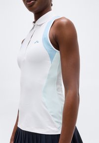 Μπλούζα πόλο σε λευκό και ανοιχτό μπλε, χωρίς μανίκια και εφαρμοστή, με φερμουάρ μπροστά, που φοράει άτομο με σκουρόχρωμη επιδερμίδα και ναυτικό πλισέ φούστα.