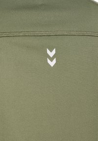 Nærbillede af olivengrønt stof med to små hvide chevroner, der peger nedad, under en vandret søm.