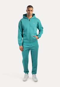 Felpa turquoise con zip abbinata a un paio di pantaloni, entrambi realizzati in tessuto morbido. Presenta una tasca a canguro e un design semplice e minimalista.
