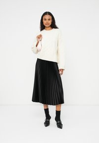 Suéter de punto trenzado color crema, falda midi plisada negra, calcetas negras y zapatos de punta con acentos metálicos. Sosteniendo un bolso de mano rosa.