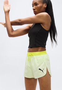 Γυναίκα που τεντώνει τα χέρια της φορώντας μαύρο αμάνικο τοπ και ανοιχτό κίτρινο σορτς Puma, μπροστά σε απλό φόντο.