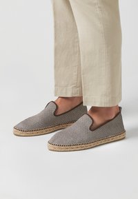 Zapatos de slip-on estilo espadrille con un patrón a cuadros marrón y blanco, suela de yute trenzado y aberturas elásticas en los laterales para facilitar su uso.