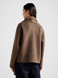 Ruda vilnos mišinio striukė su šalčio apykakle, laisvu siluetu ir nuleistais pečiais, tiesiu apatine dalimi ir subtilia tekstūra.