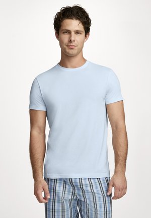 Junger Mann mit lockigem Haar, der ein hellblaues kurzärmeliges T-Shirt und blau karierte Hosen trägt, vor einem schlichten weißen Hintergrund stehend.