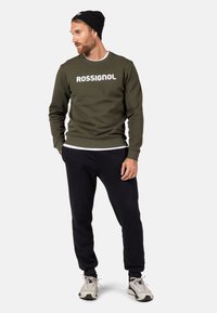 Felpa verde scuro con testo bianco "ROSSIGNOL", abbinata a pantaloni neri e scarpe da ginnastica grigie chiare. Il modello indossa un berretto nero.