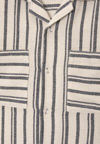 Chemise en tissu beige texturé avec des rayures noires verticales et horizontales, fermeture par boutons, col et deux poches plaquées à l'avant.
