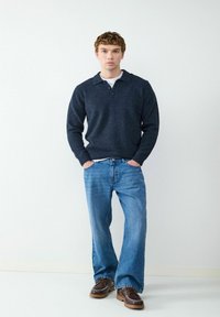 Granatowy sweter z kołnierzykiem i trzema guzikami, w zestawieniu z jasnoniebieskimi spodniami z szerokimi nogawkami oraz brązowymi butami z białymi detalami w szwie.