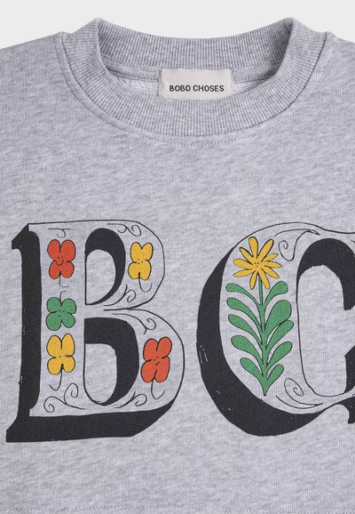 Tissu gris avec de grandes lettres décoratives "B" et "G" ornées de fleurs colorées ; étiquette de vêtement indiquant "BOBO CHOSES".
