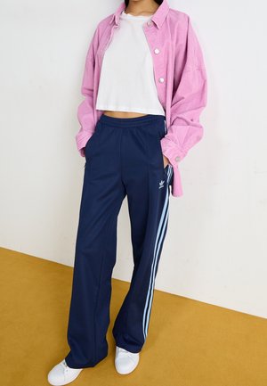 Personne portant une chemise en velours côtelé rose, un t-shirt court blanc, un pantalon de survêtement Adidas bleu marine avec des rayures bleu clair et des baskets blanches.
