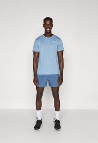 T-shirt de sport bleu clair à manches courtes avec un petit logo blanc, associé à un short bleu et des chaussures de sport noires, se tenant sur un fond uni.
