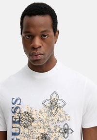 Camiseta de algodón blanca con diseños gráficos florales en azul y beige y el texto "GUESS" en el lado izquierdo. Mangas cortas y escote redondo.
